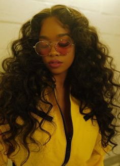 a woman with long curly hair wearing glasses and a yellow jacket is standing in front of a white brick wall