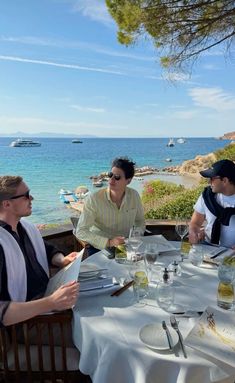 three people sitting at a table with wine glasses in front of them and the ocean behind them