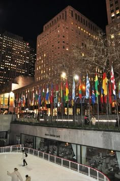 people skating on an ice rink in front of tall buildings at night with flags all around