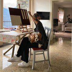 a woman sitting at a table with a dog in her lap and an easel behind her