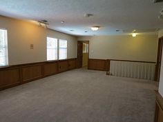 an empty living room with wood paneling and carpeted floor, windows on the far wall