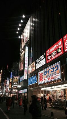 many people are walking down the street in front of some stores at night with lights on