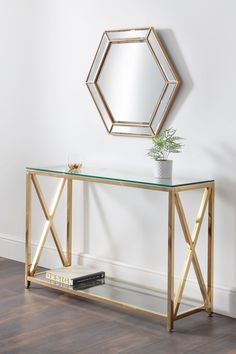 a glass and gold console table with a mirror on the wall above it, next to a plant