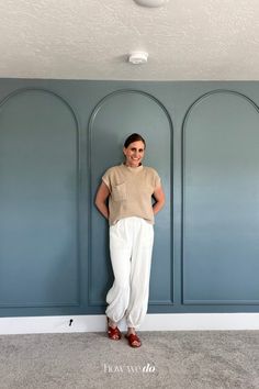 a woman leaning against a wall with her hands on her hips and wearing white pants