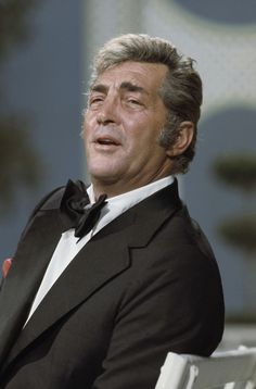 an older man in a tuxedo sitting on a white chair and looking off to the side