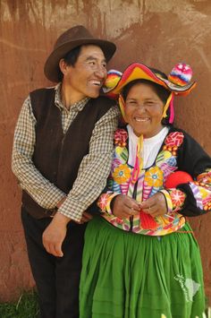 two people standing next to each other in front of a brown wall wearing colorful clothing and hats