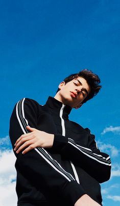 a young man is posing with his arms crossed in front of a blue sky and white clouds