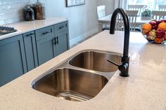 two stainless steel sinks in a kitchen with granite counter tops and blue cabinets, next to a bowl of fruit