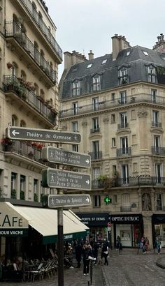 street signs pointing in different directions on the side of a road near buildings and people walking around