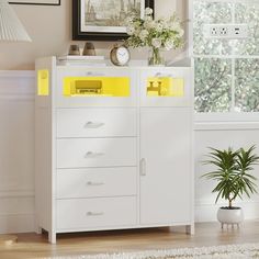 a white dresser with yellow drawers in a room next to a window and potted plant