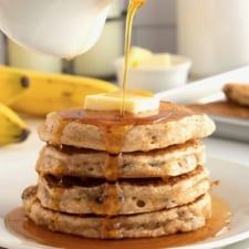 a stack of pancakes with syrup being drizzled on top and bananas in the background