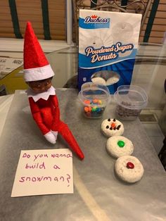 an elf sitting on top of a table next to snowman cookies and marshmallows