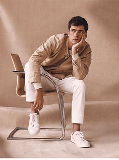 a man sitting on top of a metal chair next to a white tennis shoe in front of a beige background