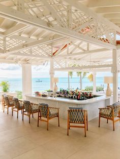 an outdoor bar with lots of chairs and bottles on the counter in front of the ocean