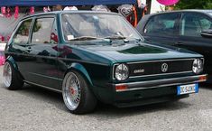 an old green car parked next to other cars in front of a tent with pink streamers