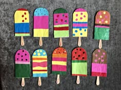 popsicles made out of colored paper are lined up on a gray surface with wooden spoons