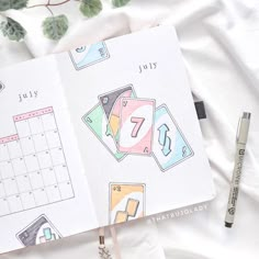 an open planner book sitting on top of a bed next to a pen and plant