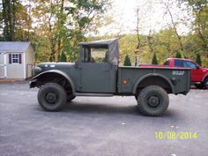 an old green truck parked in a parking lot next to a red truck and some trees