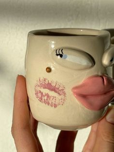 a hand holding a coffee mug with lipstick painted on it's face and tongue sticking out