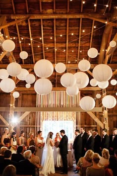 a bride and groom standing in front of their wedding party at the barn with paper lanterns hanging from the ceiling