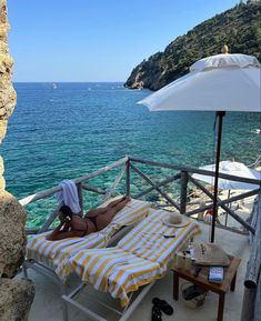 a woman laying on top of a yellow and white striped beach chair next to the ocean