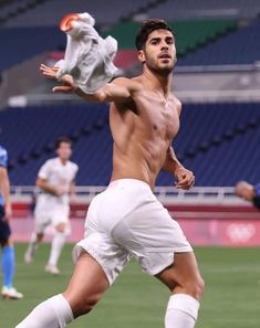 a shirtless man playing soccer on a field with other players in the stands behind him