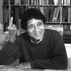 a man sitting at a table in front of a bookshelf holding up his hand