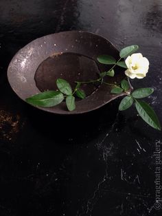 a white rose is in a black bowl on a table with some water and dirt