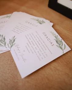 three wedding programs with green leaves on them sitting on top of a wooden table next to a black frame