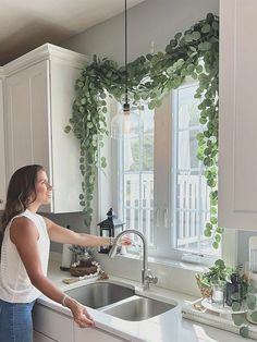 a woman standing in front of a kitchen sink next to a window filled with green plants