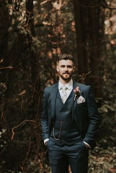 a man wearing a suit and tie standing in the woods with his hands in his pockets