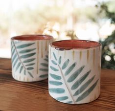 two ceramic mugs sitting on top of a wooden table next to a plant pot