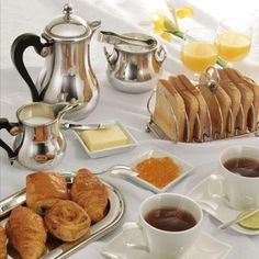 a table topped with plates of food and cups filled with liquid next to silver teapots