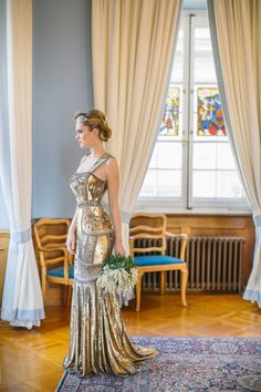 a woman standing in front of a window wearing a gold dress and holding a bouquet