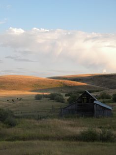 an old barn sits in the middle of a grassy field with rolling hills behind it