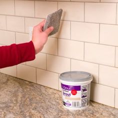 a person holding a cloth over a can of paint on a counter top next to a white tile wall