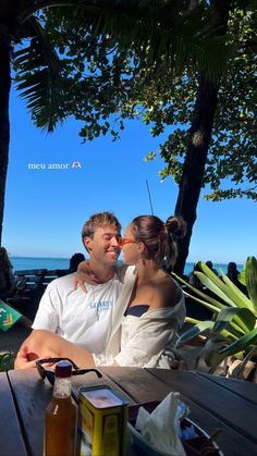 a man and woman sitting next to each other at a table near the ocean with drinks in front of them