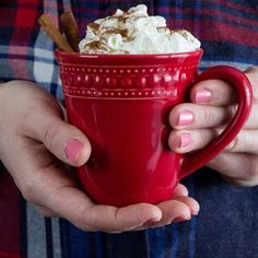 a woman holding a red cup with whipped cream and cinnamon sticks in it's hands