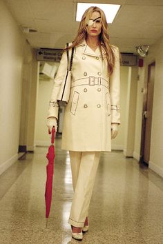 a woman with an umbrella walks down a hallway in a trench coat and matching heels