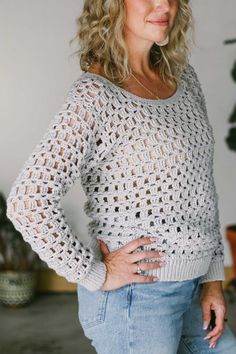 a woman standing in front of a plant wearing a white crochet sweater and jeans