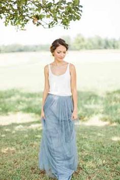 a woman standing under a tree wearing a blue skirt and white tank top with her hands in her pockets