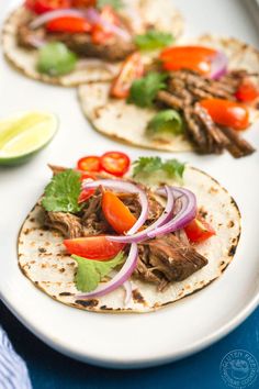 three tacos with meat, onions and peppers on a white plate next to lime wedges