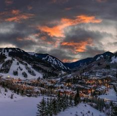 the sun is setting over a snowy mountain town with trees and mountains in the background