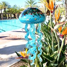 a blue glass jellyfish hanging from a plant next to a swimming pool with flowers in the foreground