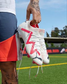 a person with tattoos holding a pair of white and red shoes on the grass at a soccer field