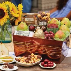 a wooden box filled with assorted fruit and snacks next to a vase full of sunflowers