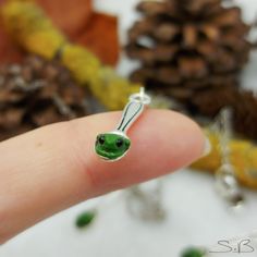 a tiny green animal charm sitting on top of a persons finger in front of pine cones