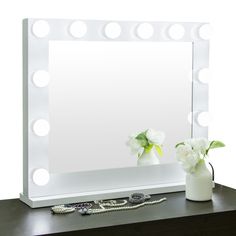 a white vase with flowers sitting on top of a table next to a lighted mirror