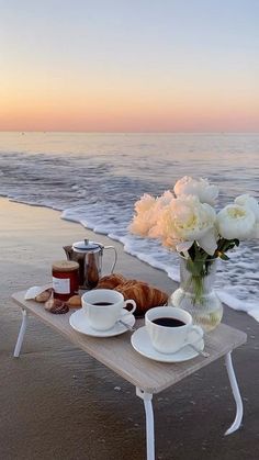 two cups of coffee are sitting on a table near the beach with flowers and croissants