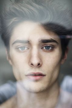 a man with blue eyes looking at the camera through a glass window, his hair is blowing in the wind